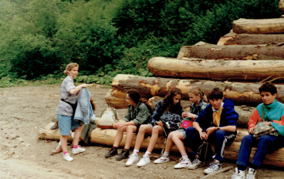 Dzieci siedzą na balach drewna. Obok opiekunka.