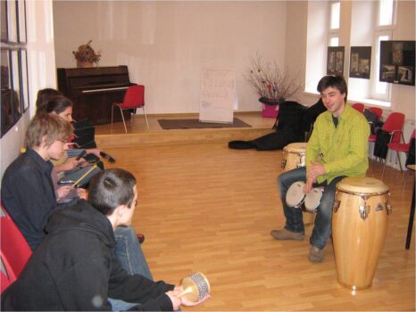 Grupa młodzieży w sali widowiskowej MDK. Słuchają jak chłopak gra na bębnie.