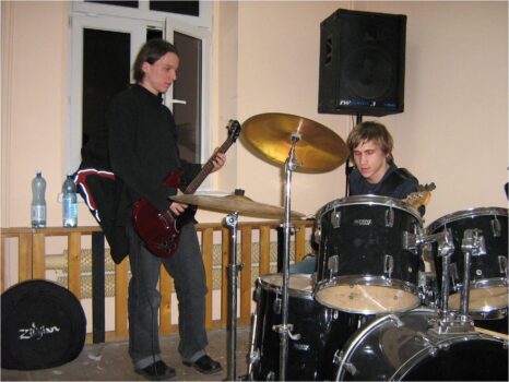 Młodzież na próbie muzycznej. Jeden chłopak siedzi przy perkusji, drugi gra na gitarze. 