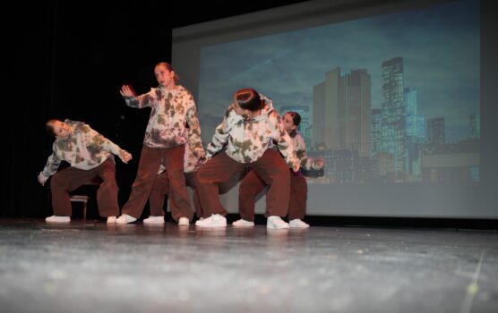 Grupa tancerek hip-hop w bluzach i brązowych spodniach tańczy na scenie. Stoją w różnych dziwacznych pozycjach.