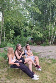 Trzy dziewczyny siedzą na ławce w ogrodzie. W tle uroczy zagajnik.
