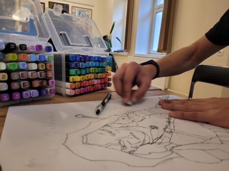 Widok na pracę wykonywaną ołówkiem, w tyle kolorowe pisaki i kredki, widać też dłoń, która rysuje.