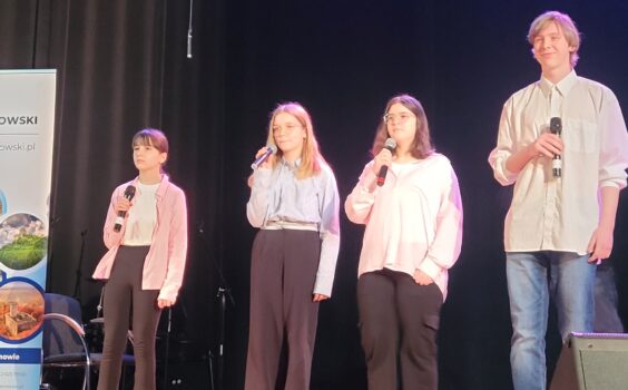 Trzy dziewczyny i jeden chłopak stoją na scenie- w ręku mają mikrofony.