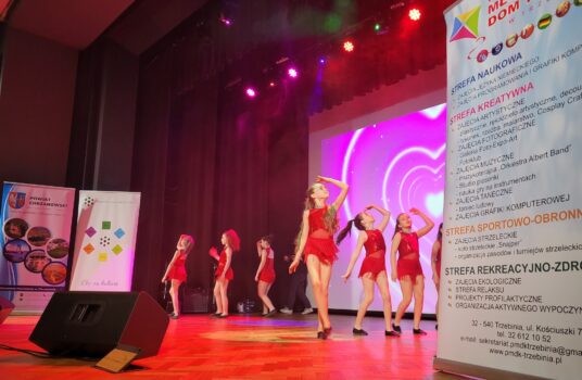 Widok na scenę LCK oświetloną czerwonym światłem. Na scenie grupa młodych tancerek w czerwonych sukienkach. Dziewczyny tańczą. W pierwszym planie widać fragment rollupu PMDK Z tyłu - LCK i starostwa.