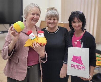 Trzy uśmiechnie.te kobiety. Jedna trzyma torbę z różowym logo PMDK, środkowa ma czarną sukienkę, trzecia trzyma w ręku piłeczkę antystresową i dwa kurczaczki uszyte w PMDK.